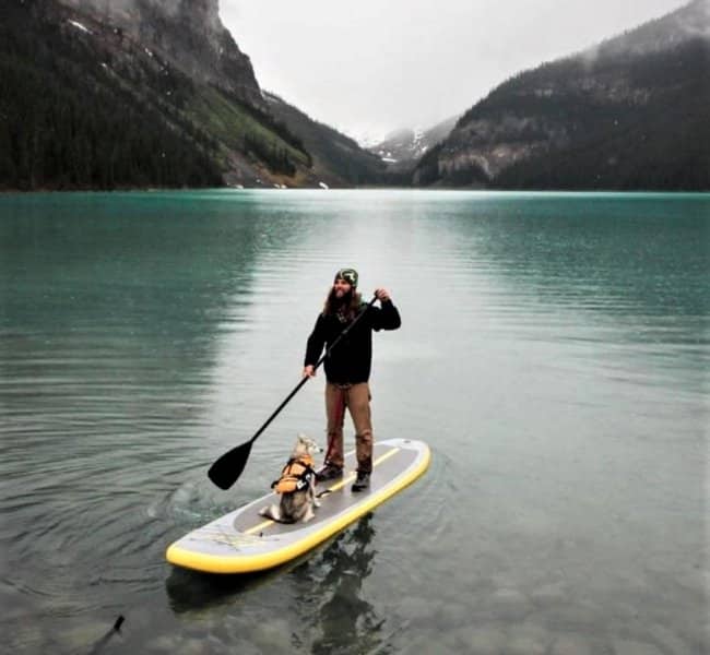man standing on a kayak with his dog in a lake