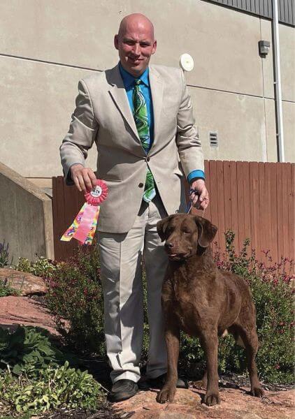 Owner-handler in a suit and tie holding a ribbon with a brown dog, showcasing elegance and companionship.